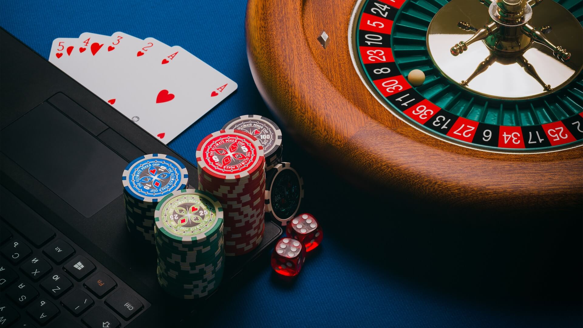 A laptop next to a roulette wheel, surrounded by poker chips, dice, and playing cards