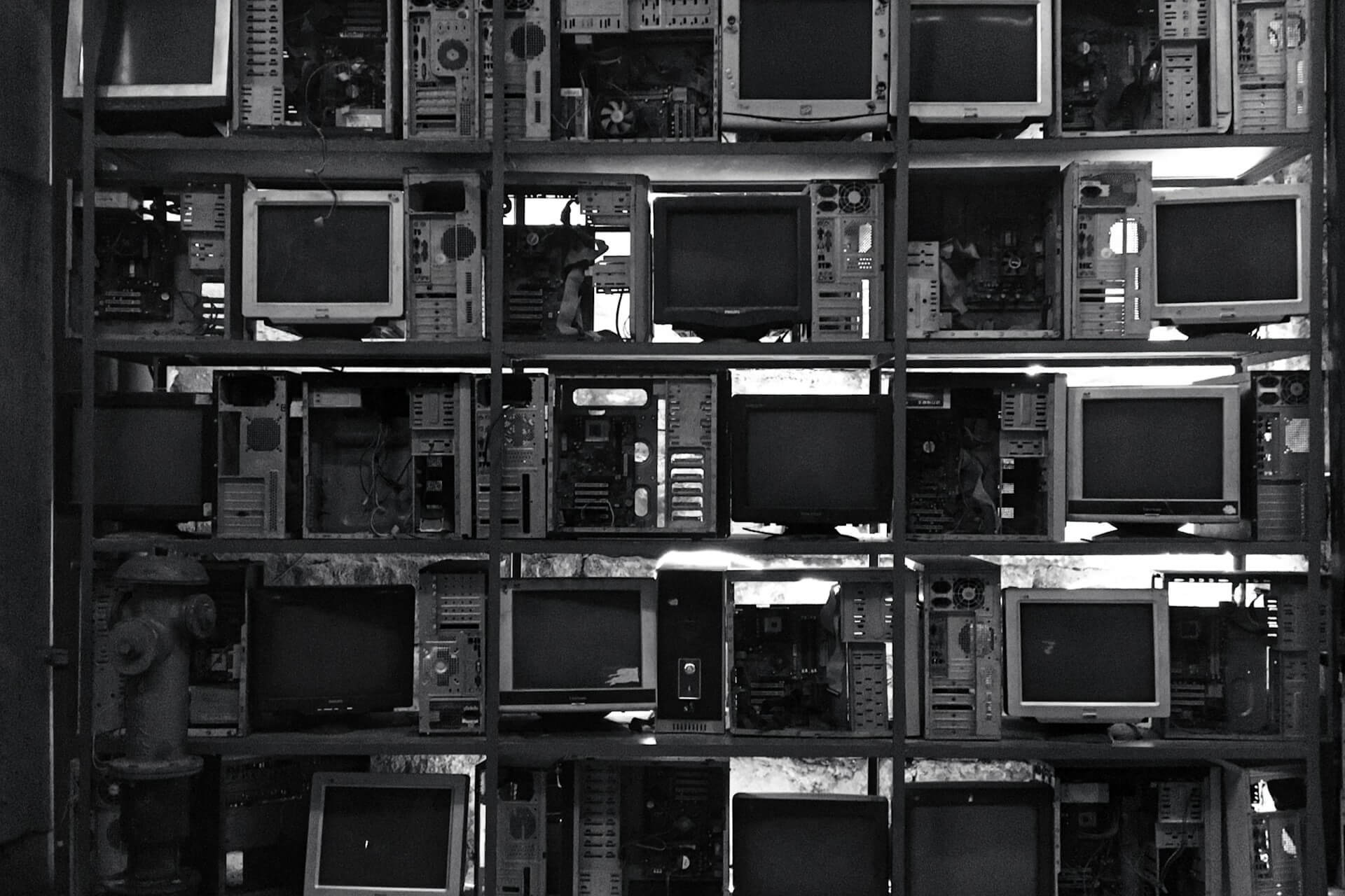 Black-and-white photograph of shelves loaded with broken desktop computers and monitors