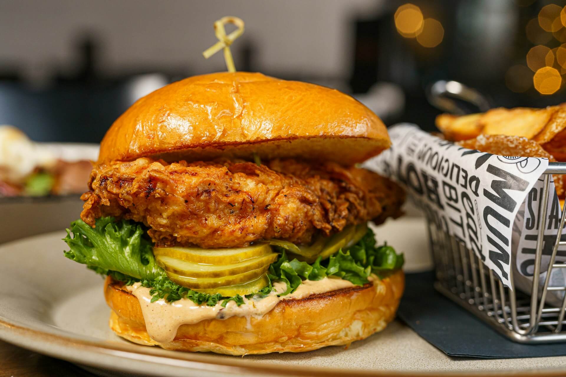 A well-crafted chicken sandwich with pickles, lettuce, and sauce, served next to a basket of French fries