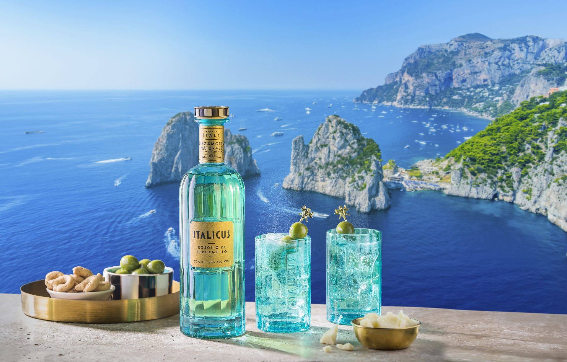 A bottle of ITALICUS Rosolio di Bergamotto and two cocktails, resting on a ledge overlooking the ocean.