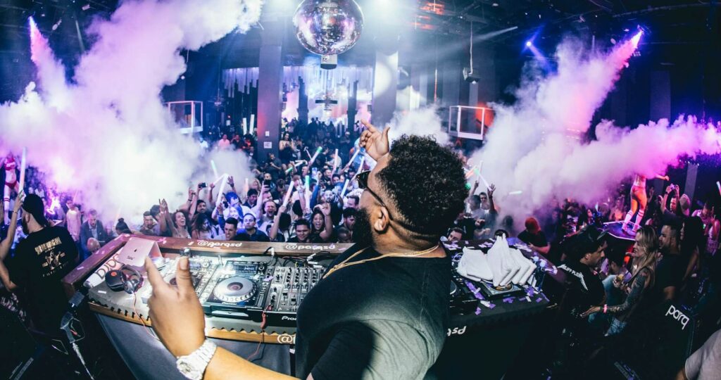 A DJ performing inside a nightclub, with the camera over his shoulder looking into the crowd