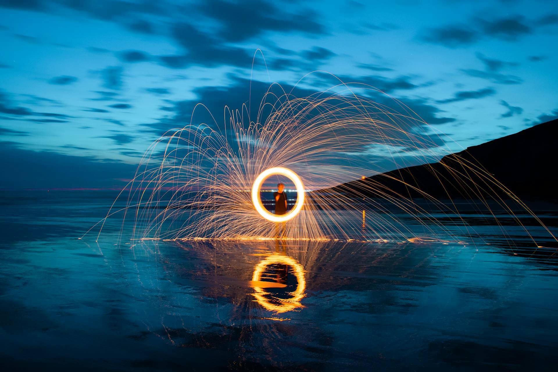 A person standing in water, creating a circle and sparks with steel wool and fire