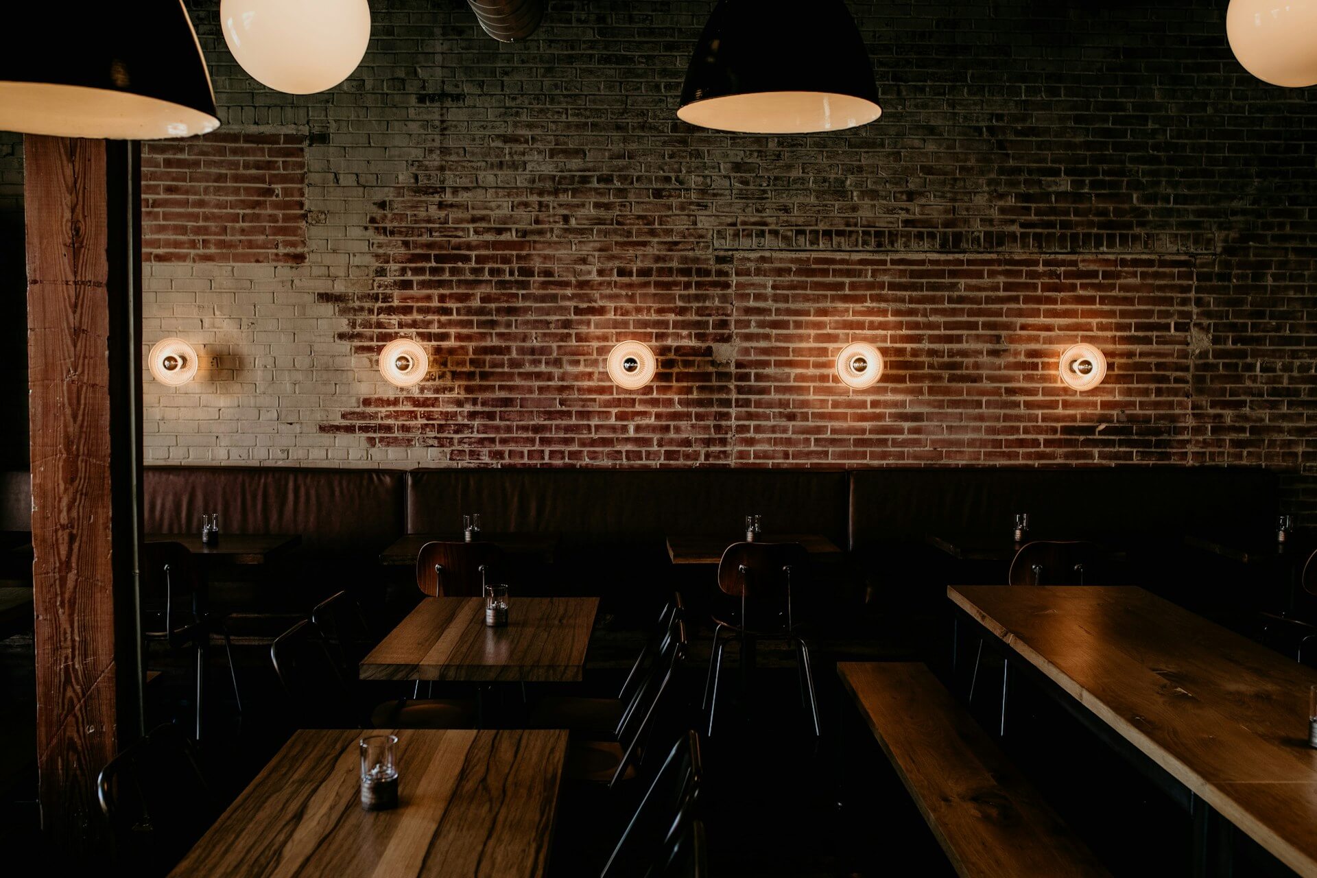 Empty restaurant dining room with wooden tables, dark booth seating, exposed brick walls, and warm pendant lighting.